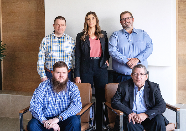 Executive Team Group Photo A group portrait of five professionals in a modern office. Two men are seated in leather armchairs in the foreground, while two men and one woman stand behind them. They are dressed in business-casual attire including blazers and button-down shirts.