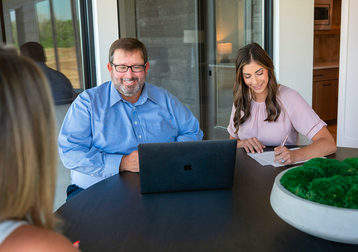 Personalized Client Meeting A man in a blue shirt and a woman in a pink blouse sitting at a dark table with a client. The man is smiling toward a laptop, while the woman is writing on a document. A bowl of decorative green moss is in the foreground.